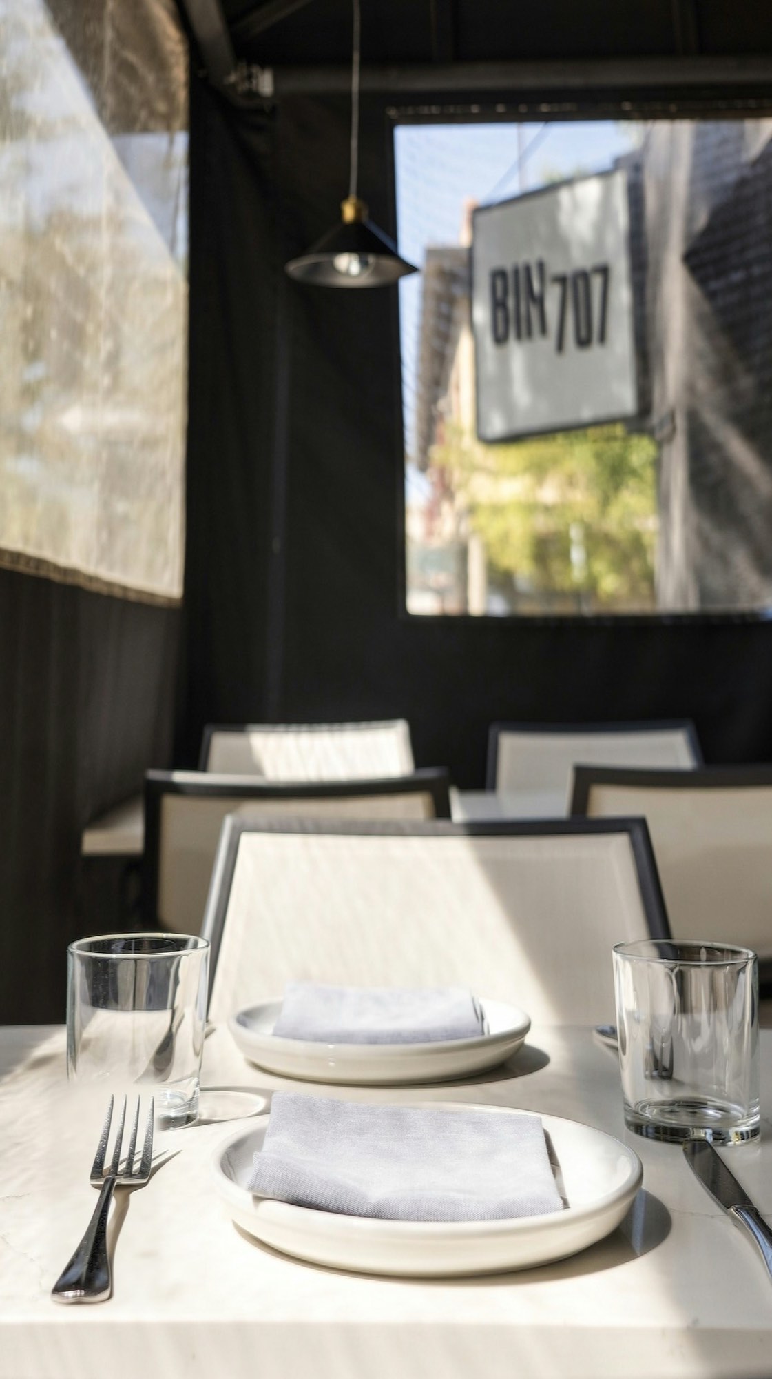 A set table inside the Bin 707 restaurant patio enclosure in Grand Junction, with the restaurant sign visible through a clear window panel