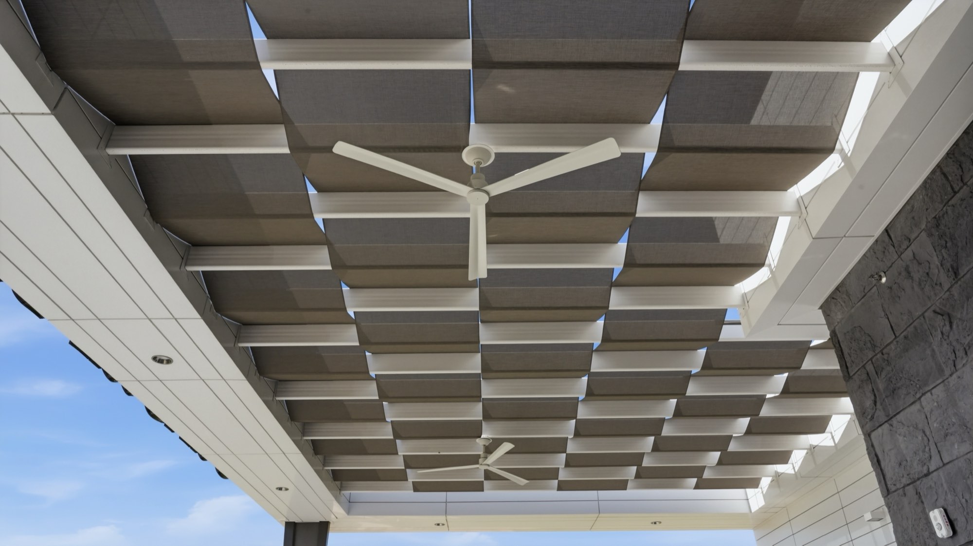 Looking up at a grid of tensioned fabric shade panels and ceiling fans under a building overhang
