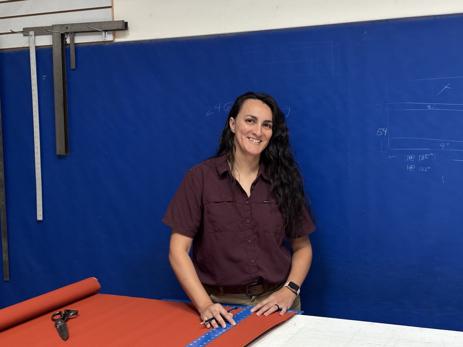 Madison Urrutia, owner of Canvas Shade, at the cutting table in the workshop with a roll of orange fabric and a chalk-marked blue wall behind her